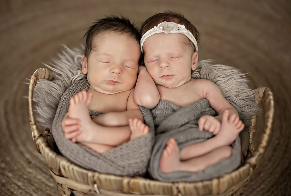 Two newborn babies sleeping in a basket wearing grey blankets