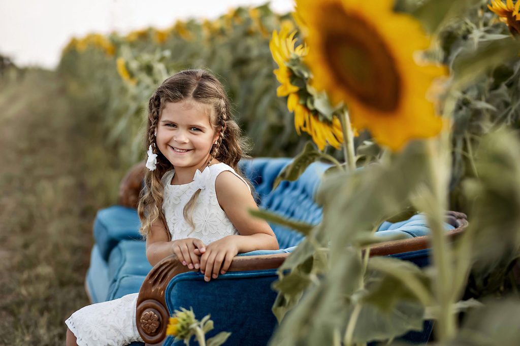 Smiling girl in white dress on blue couch among sunflowers